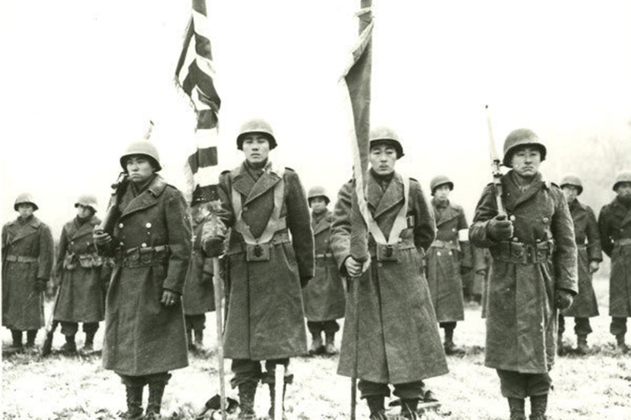 Four service members stand at attention, two of whom hold flags over their shoulders. Others stand in line behind them.