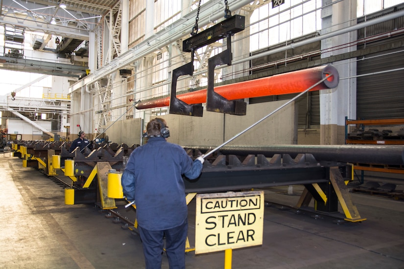 A man guides a metal barrel suspended by a crane inside of a factory.