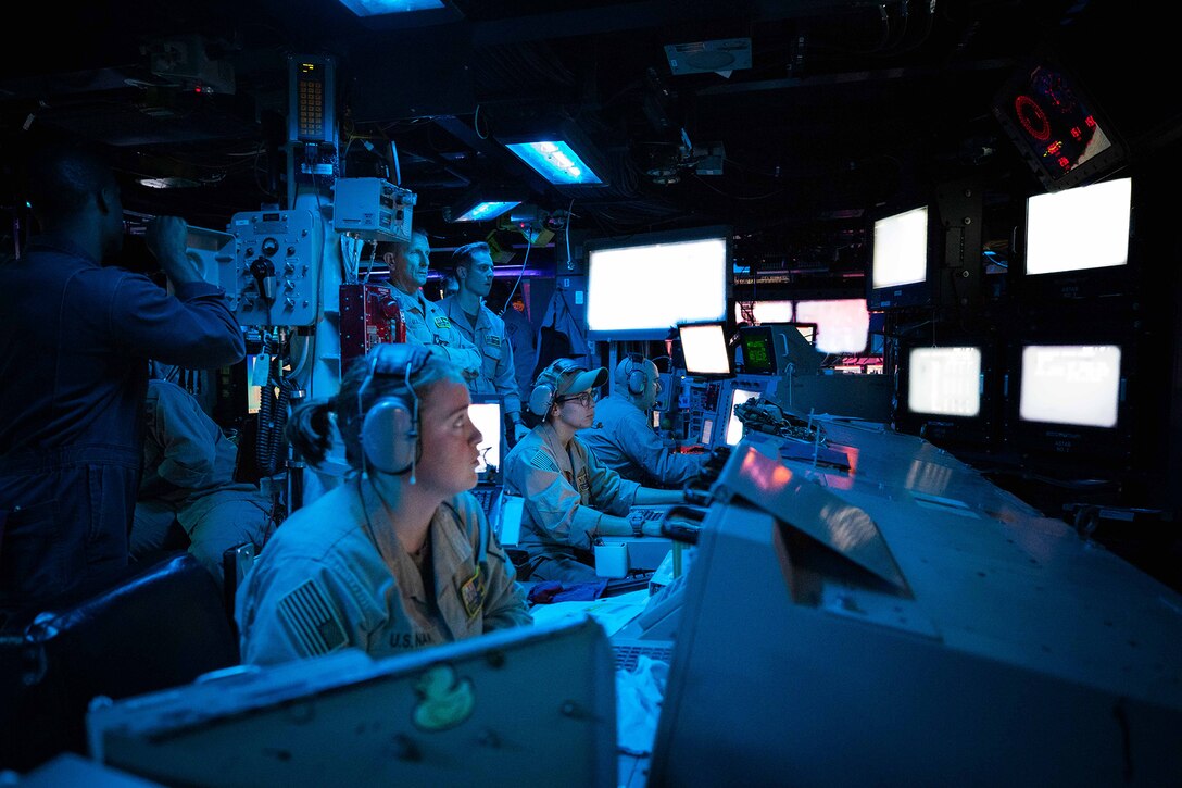 Three sailors sit and look at multiple monitors in a dark room while several more stand behind them.