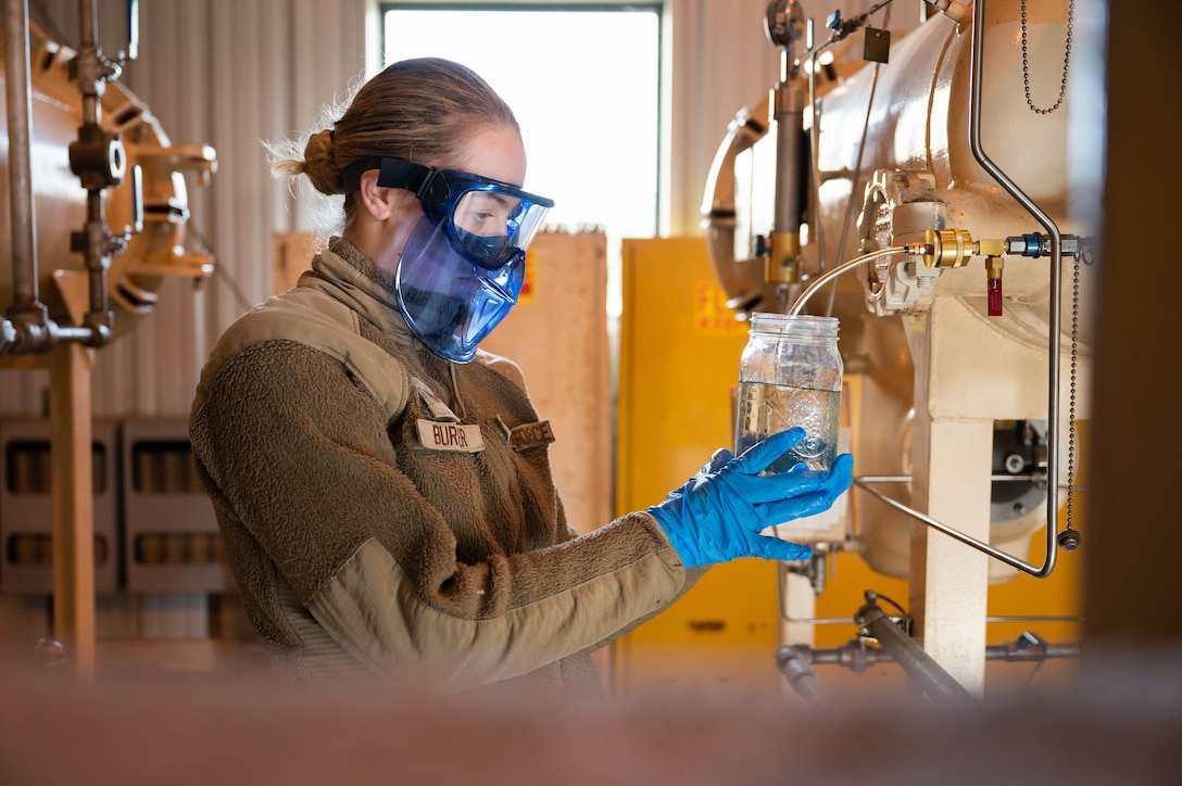 A uniformed airman wearing a protective eye and face shield holds a jar of fuel.