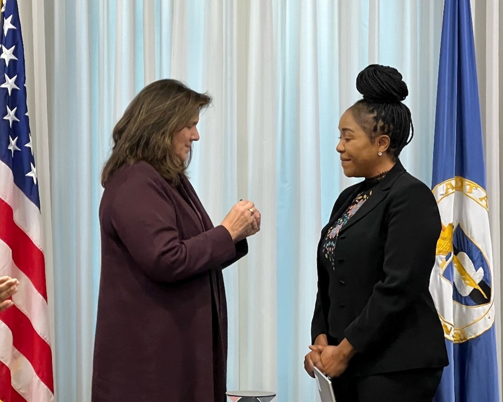 Two business professionals standing in front of a pearlescent backdrop. A United States of America flag borders the left side and the DCAA flag borders the right side of the frame.