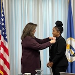 Two business professionals standing in front of a pearlescent backdrop. A United States of America flag borders the left side and the DCAA flag borders the right side of the frame.