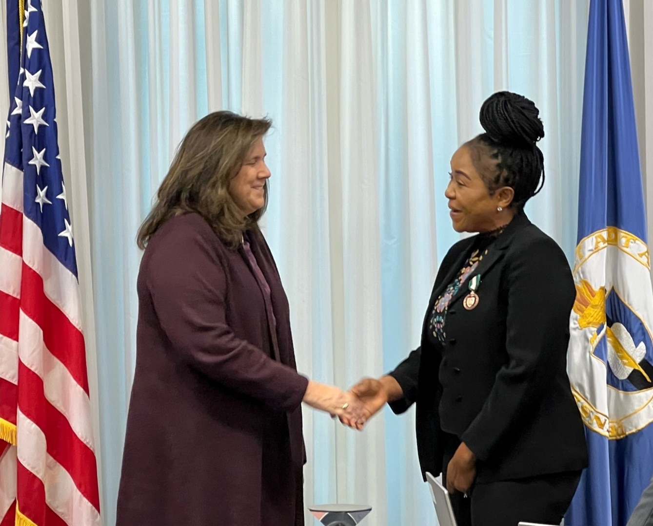 Two business professionals standing in front of a pearlescent backdrop. A United States of America flag borders the left side and the DCAA flag borders the right side of the frame.