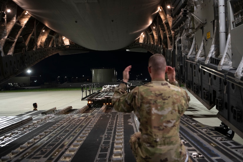 A man in uniform signals with his hands on the ramp of a military cargo plane.
