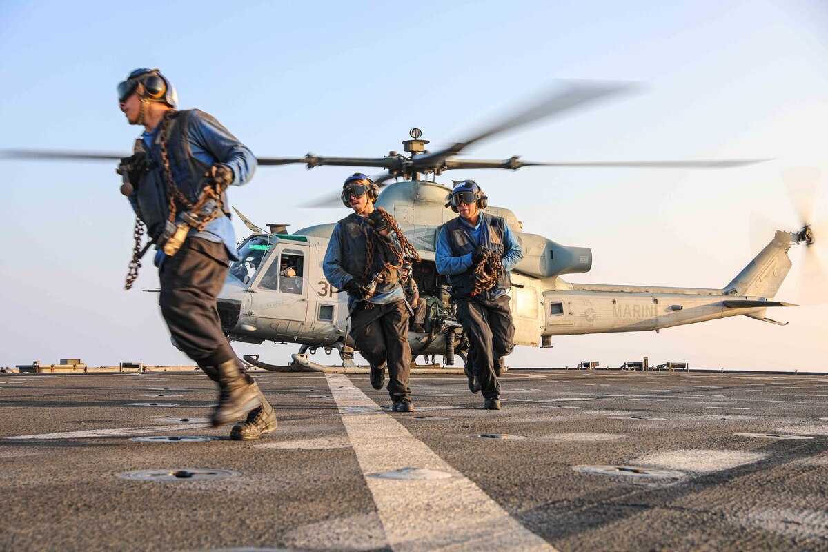 Three sailors in military uniform perform flight operations. A helicopter is behind them.