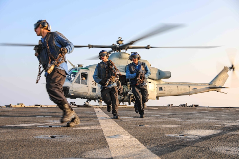 Three sailors in military uniform perform flight operations. A helicopter is behind them.