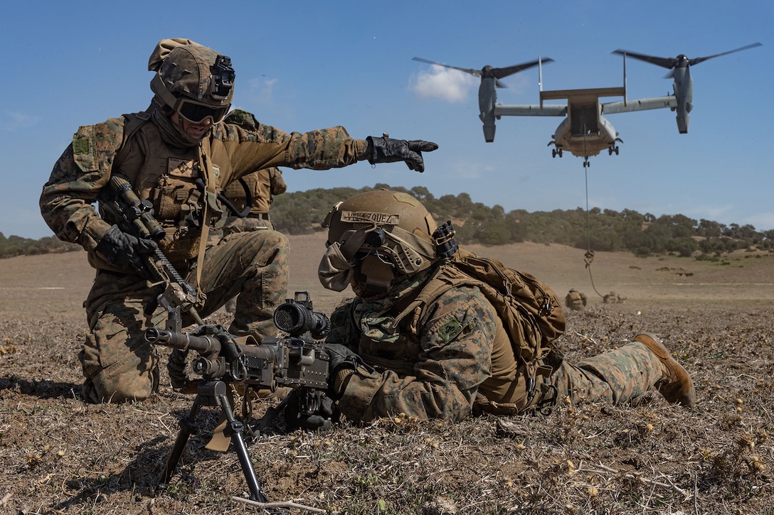 One soldier lays on the ground with a weapon while another soldier points and gives direction.