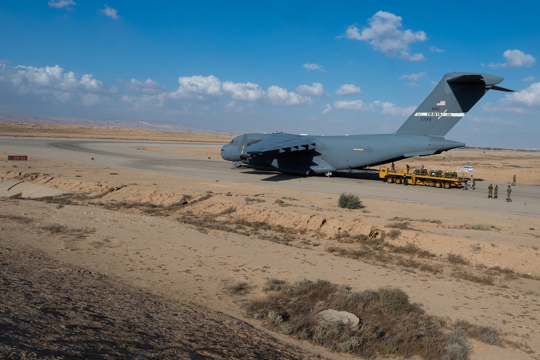 A large aircraft sits on a desert runway with service members standing behind it.