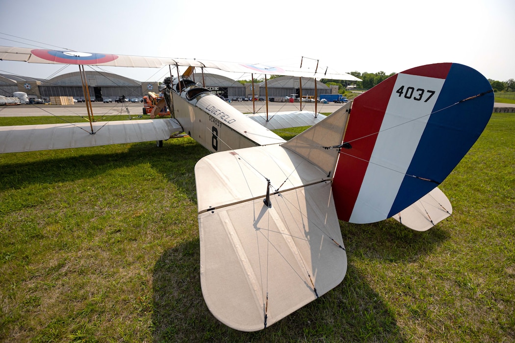 The Jenny on great grass from behind left side. the tan airframe is visible with the photo focusing on the red, white and blue vertical stripe on the tail with the number 4037.