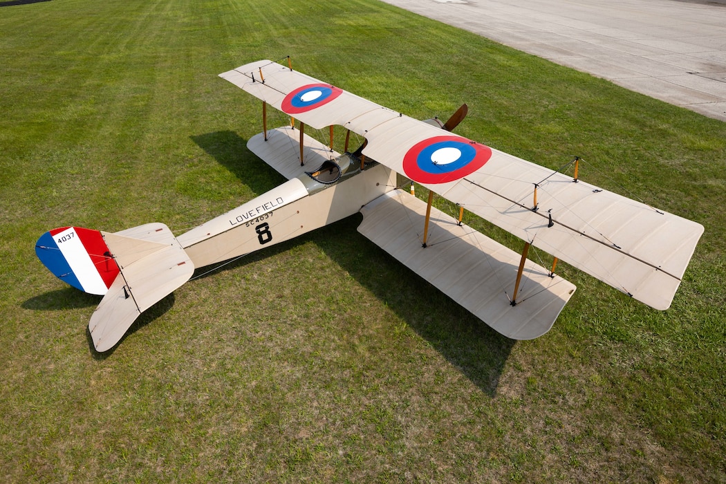 Overhead image of the Jenny in the grass outside the restoration hangar. From this view you can see the tan aircraft coloring with the red, blue and white circle insignia on the top and the red, white and blue stripes on the tail.