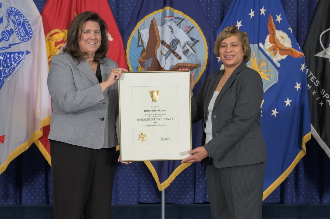 Two career professionals hold an award in front of a background of flags.