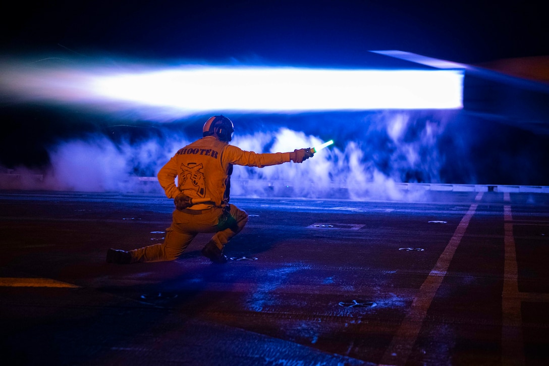 A sailor holding a flashlight signals an aircraft’s launch from a ship at night.