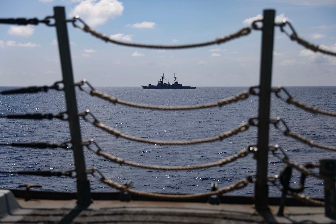A ship sails in the distance. The ship can be seen through a rope and metal railing.