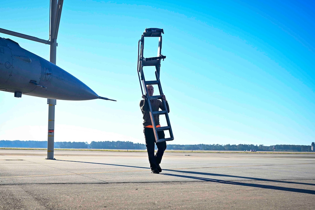 An airman carries a ladder next to a parked aircraft.