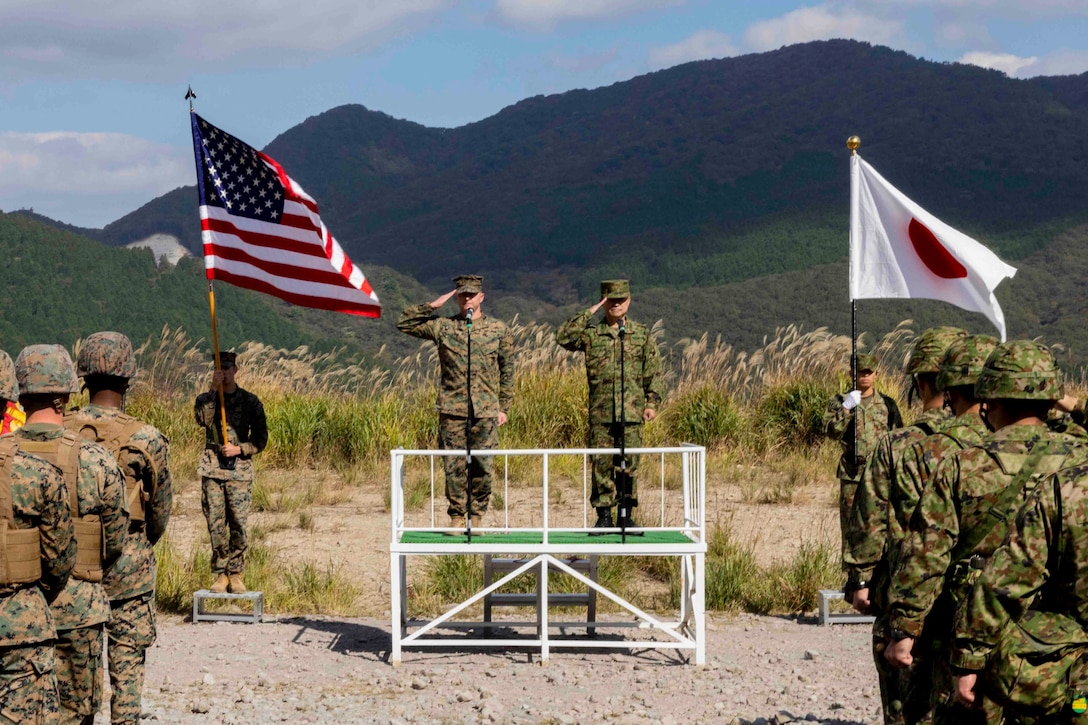 A Marine and a foreign service member salute while standing on stage between two service members holding flags in front of troops standing in formation.