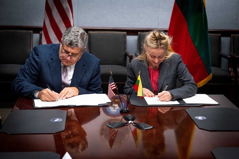Two people in business attire sign documents at a wooden table with flags behind them.