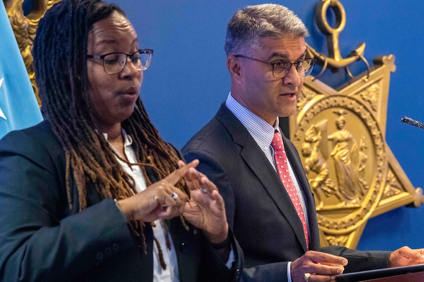 A woman signs with her hands as a man speaks at a lectern.