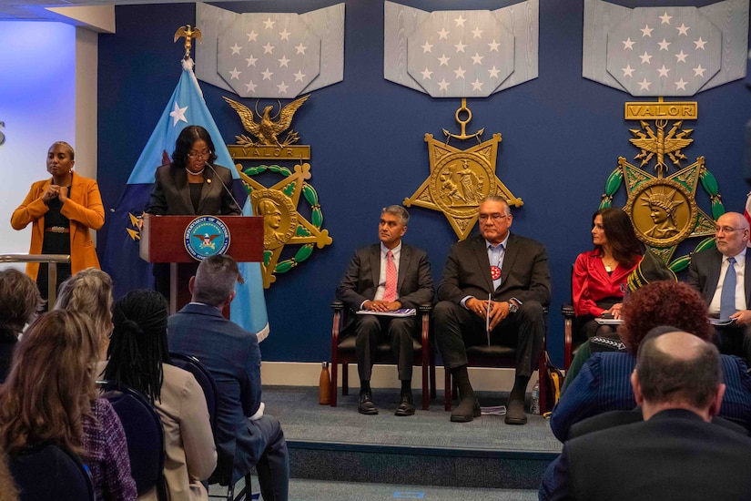 A woman in civilian attire stands at a lectern with others seated behind her.