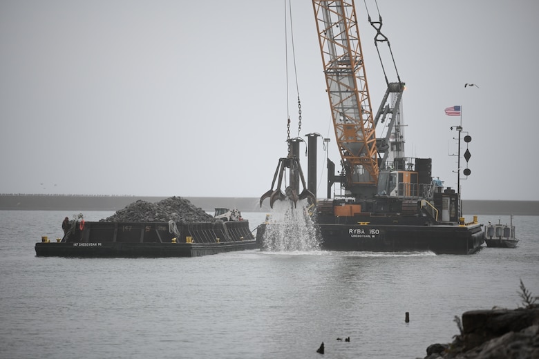 A crane on a derrickboat lifts a mechanical claw out of water next to a barge full of stones.