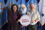 Two professionally dressed people hold a sign in front of flags.