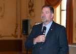 Director Mike Cannon, wearing a blue jacket holds a mic while making his opening remarks. the room he is in has tan walls with a large window behind him the walls have painted murals but are not discernable due to be out of focus from shallow depth of field used to photograph Cannon.