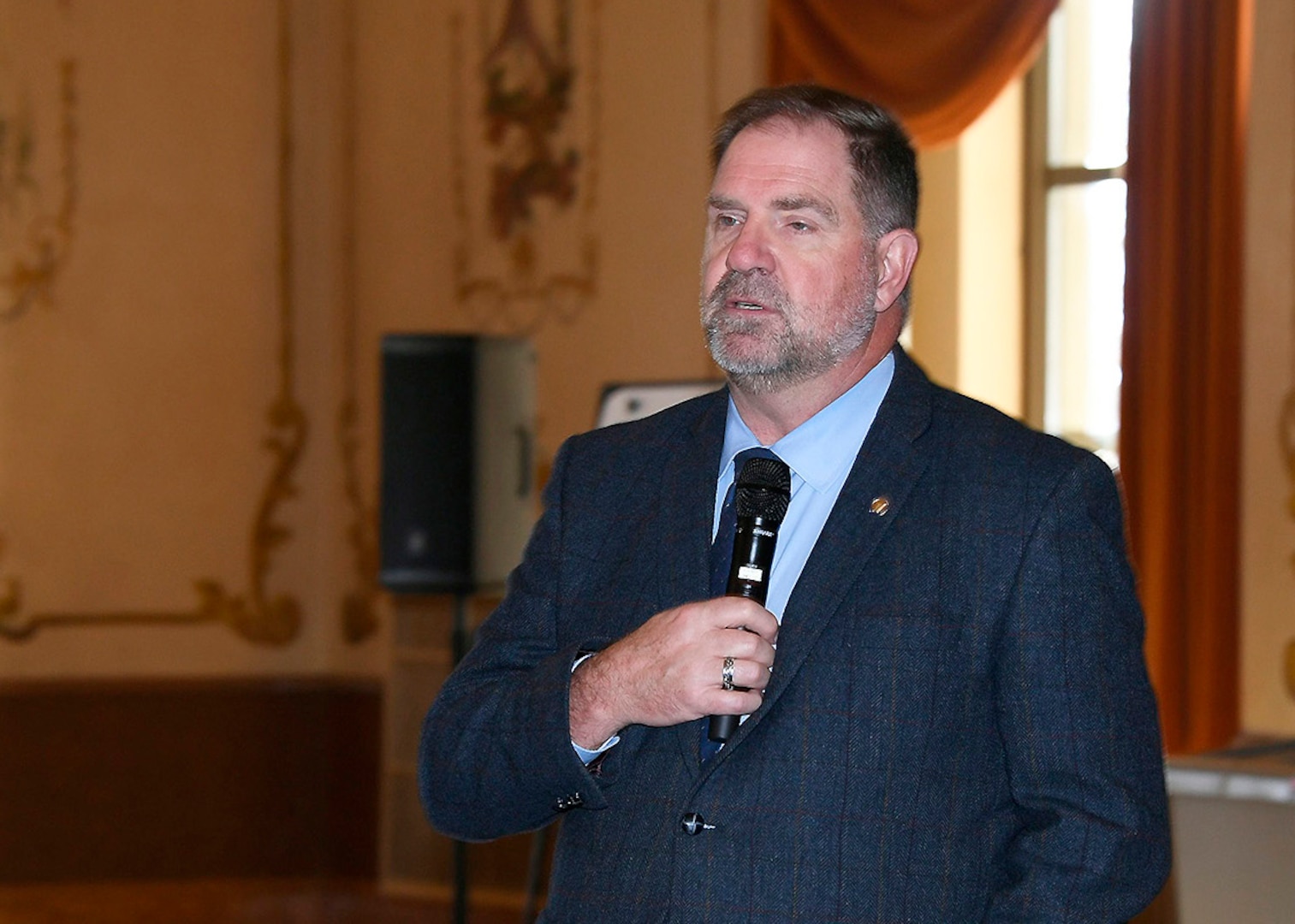 Director Mike Cannon, wearing a blue jacket holds a mic while making his opening remarks. the room he is in has tan walls with a large window behind him the walls have painted murals but are not discernable due to be out of focus from shallow depth of field used to photograph Cannon.
