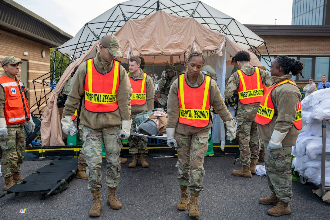 Airmen carry a dummy on a gurney out of a makeshift tent in front of a building as fellow airmen watch.