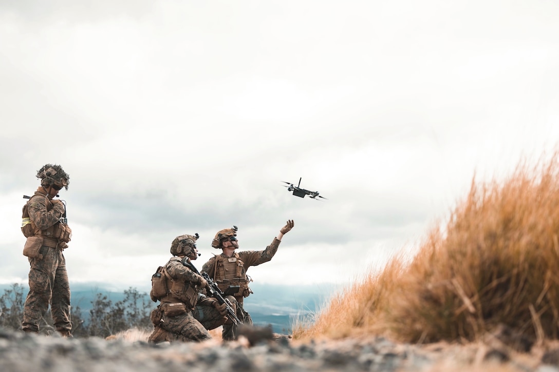 A Marine stands watch as two Marines kneeling on a rocky surface release a drone into a cloudy sky with a patch of dry grass to the right.