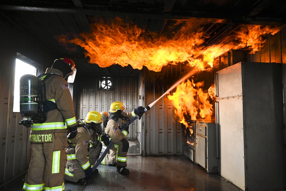 An instructor watches as two kneeling airmen use a hose to put out a simulated kitchen and ceiling fire.