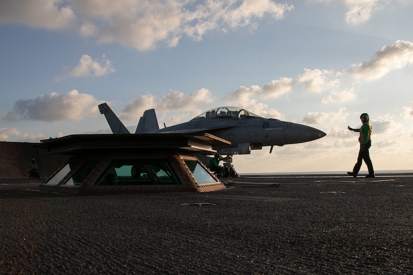 A man in flight deck equipment signals with his hands to a pilot in the cockpit of a Navy fighter jet.