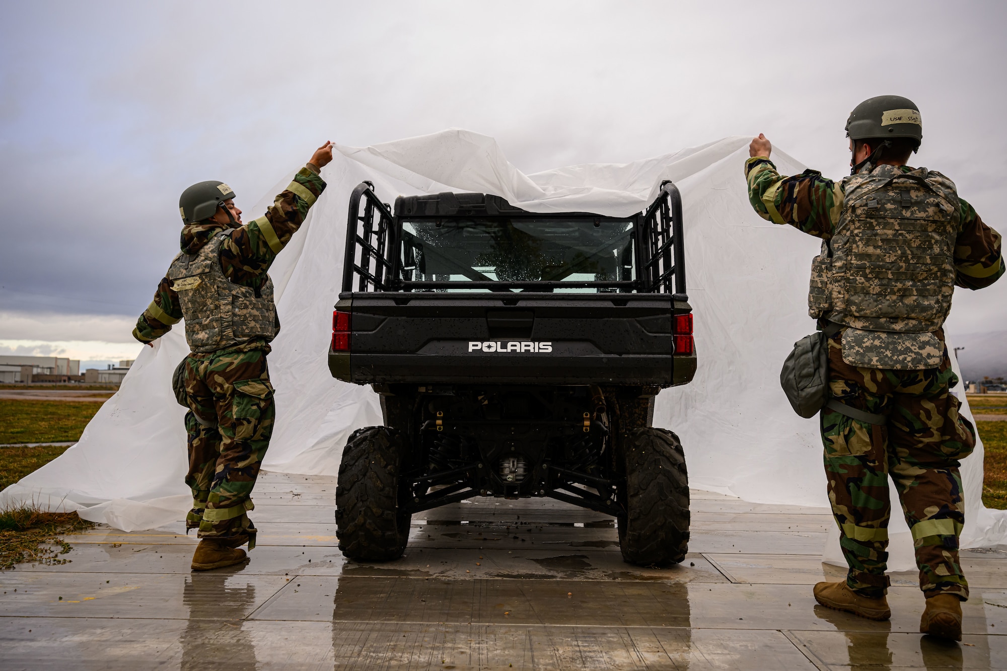 Two Airmen in chem gear cover a utility vehicle with opaque plastic