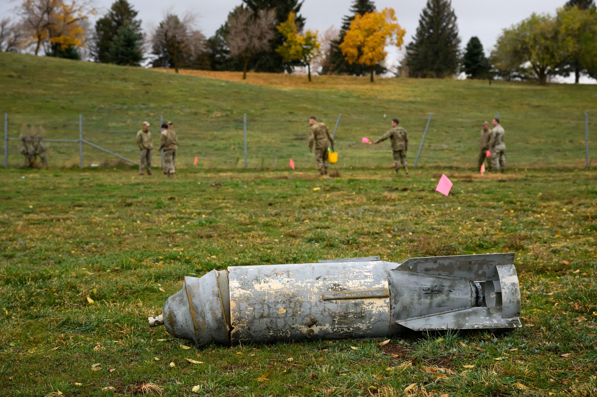 A dummy bomb lies in the foreground while Airmen walk in a field.