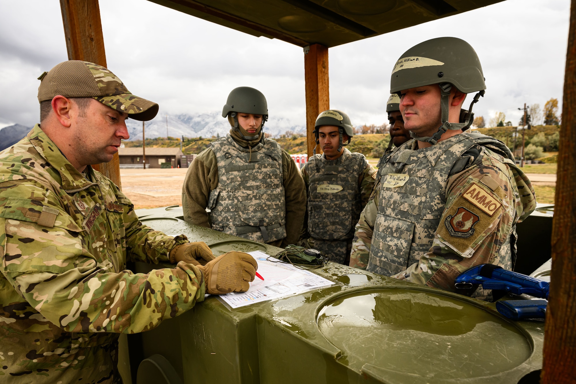 An instructor uses a map to teach three Airmen all standing in an outdoor shelter