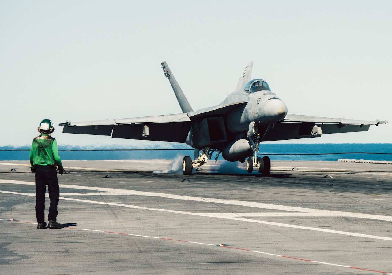 A military jet lands on the deck of an aircraft carrier during the day as a person in a military uniform observes.