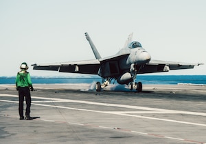 A military jet lands on the deck of an aircraft carrier during the day as a person in a military uniform observes.