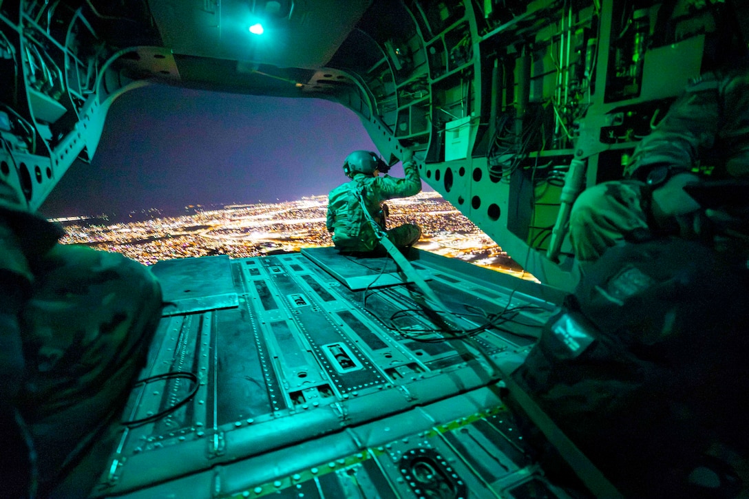 A guardsman sits at the back of an airborne aircraft looking out over a city night.