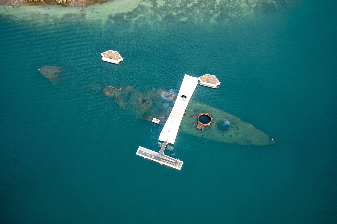 Aerial view of a long floating white structure over wreckage from a ship in teal water.