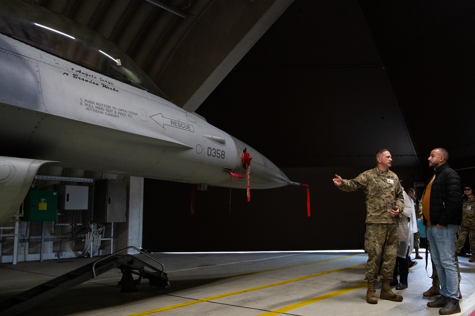 A U.S. Airman standing in a hangar gestures towards a fighter aircraft behind him as he faces a civilian who is looking at the aircraft.
