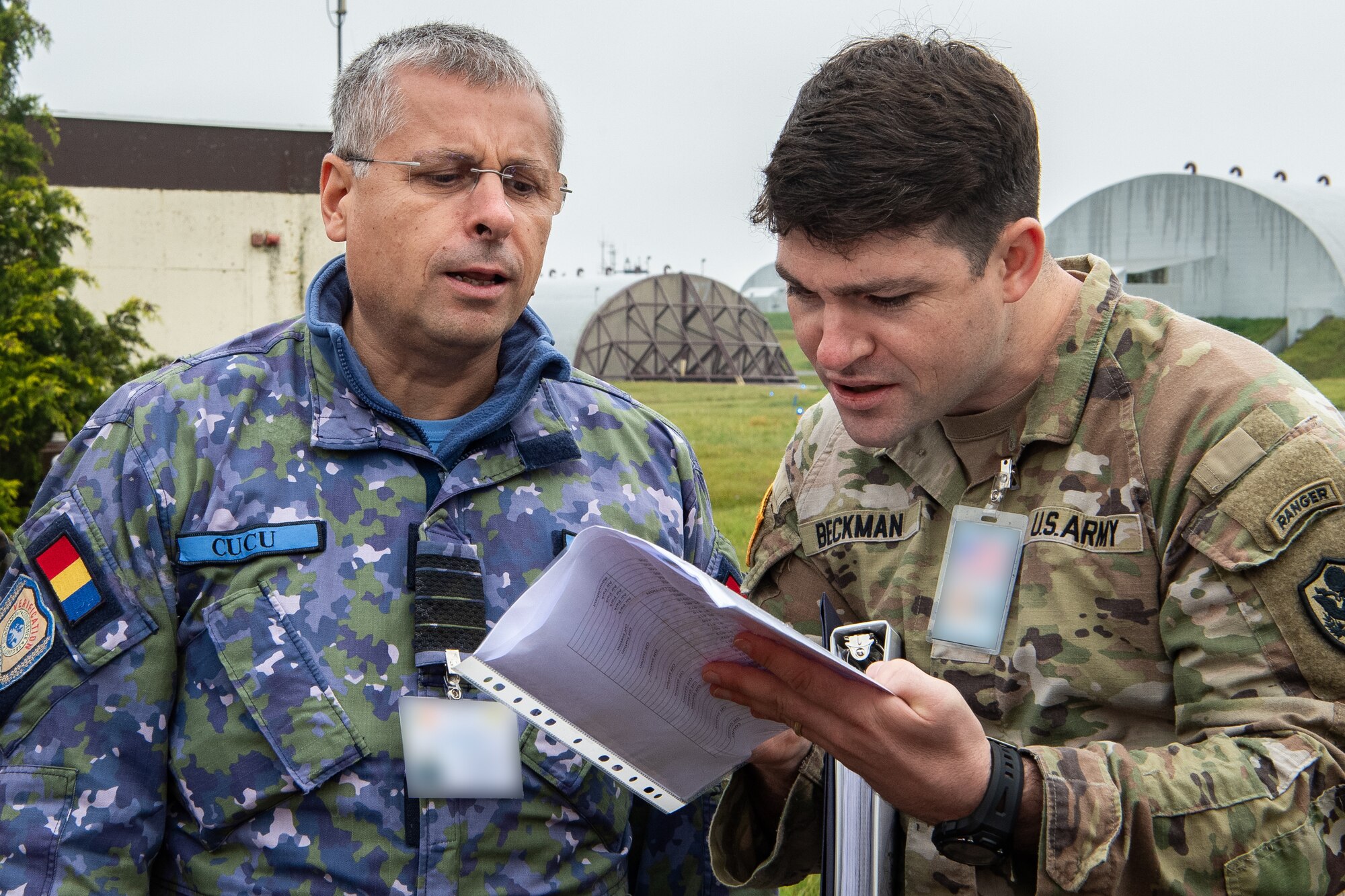 A Romanian Airman and a U.S. Army Soldier look at a paper in a plastic sheet. Aircraft hangars are seen in the background.