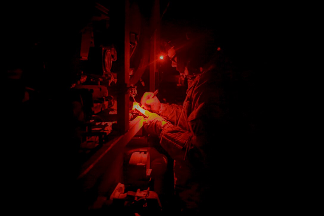 A soldier wearing a headlamp writes notes while standing in front of a mechanical system.