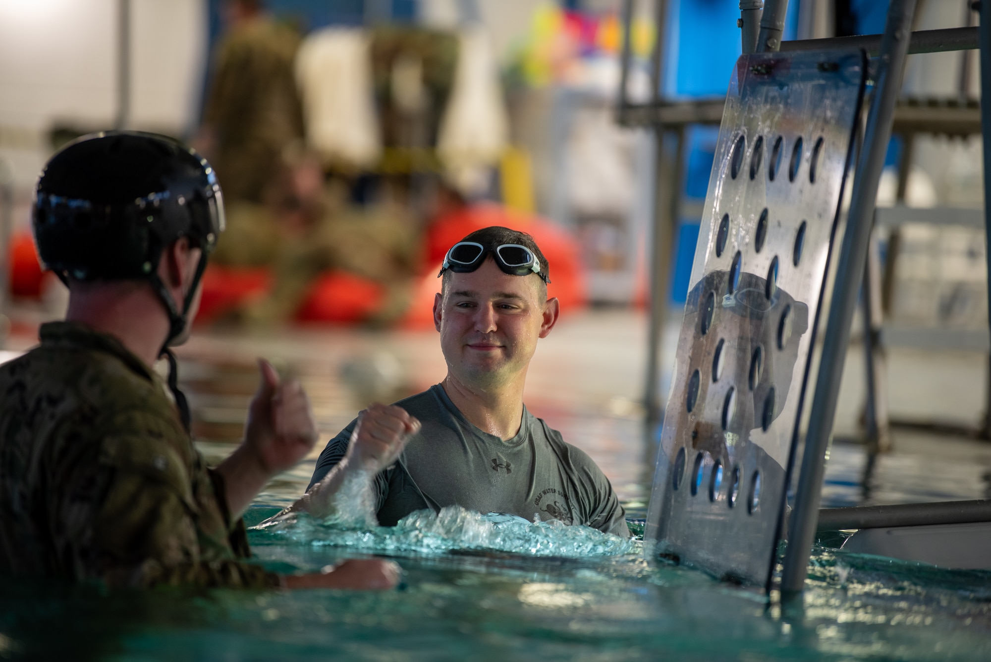 A instructor wearing a grey shirt and swimming goggles on his forehead fist bumps a soldier wearing a black helmet. Both of them are in a pool and treading water by a metal structure.