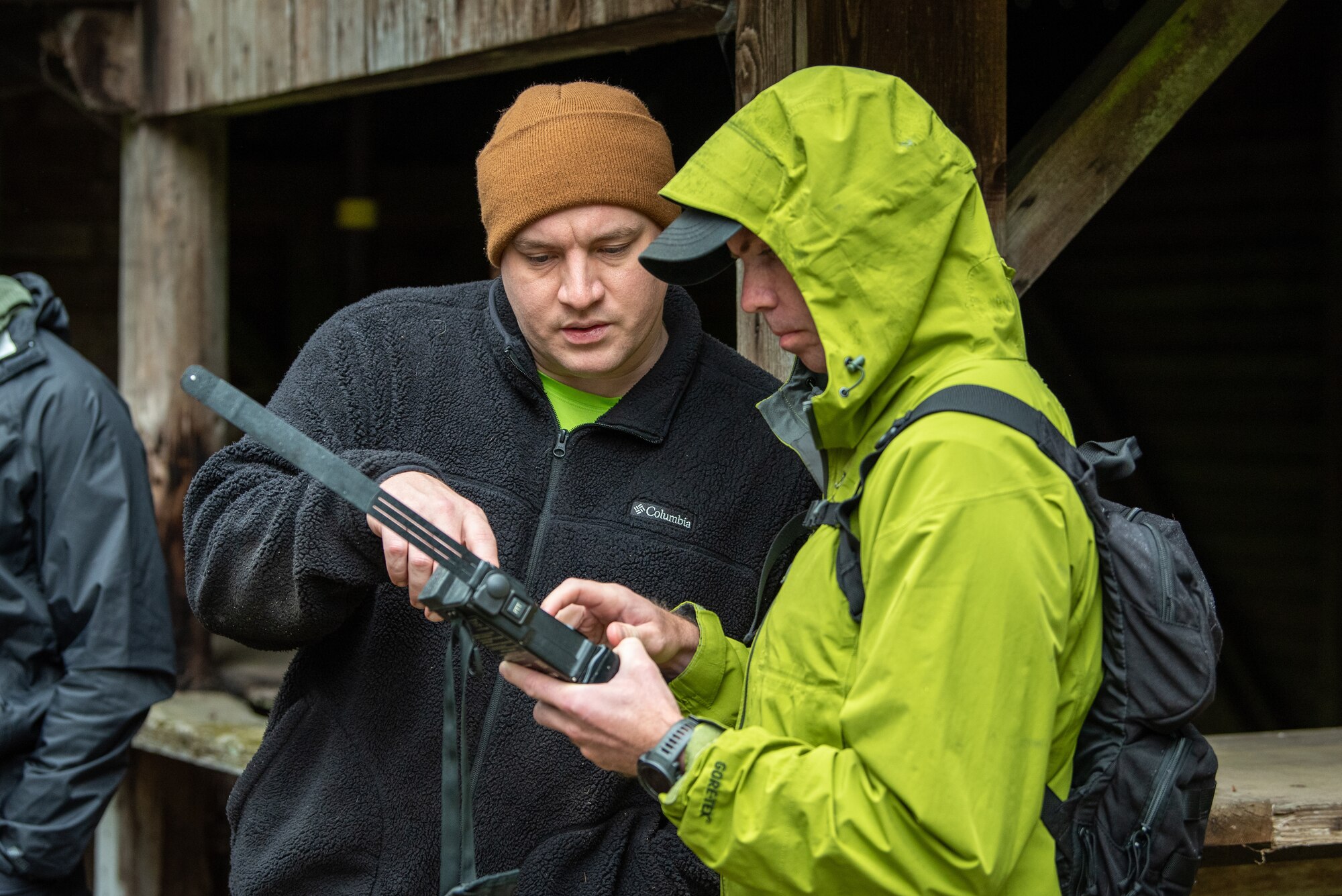 A man in a black fleece jacket and tan beanie points at military walkie talkie while talking to a man holding the walkie talkie while wearing a ball cap, a light green rain jacket with the hood up, and a small black backpack.
