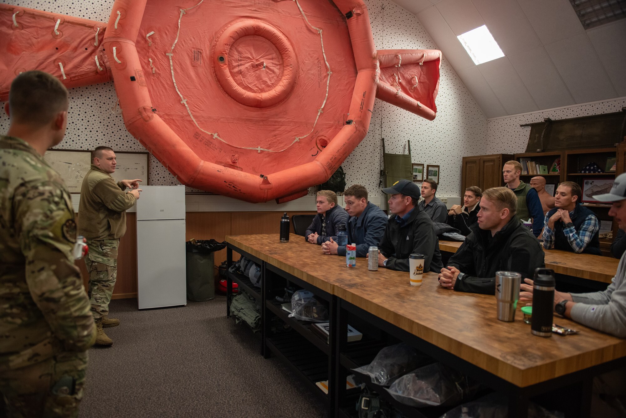 Two Airmen in uniform talk to two rows of students in civilian clothes sitting at long wooden tables. A giant inflatable lift raft is hung on the wall as a decoration.