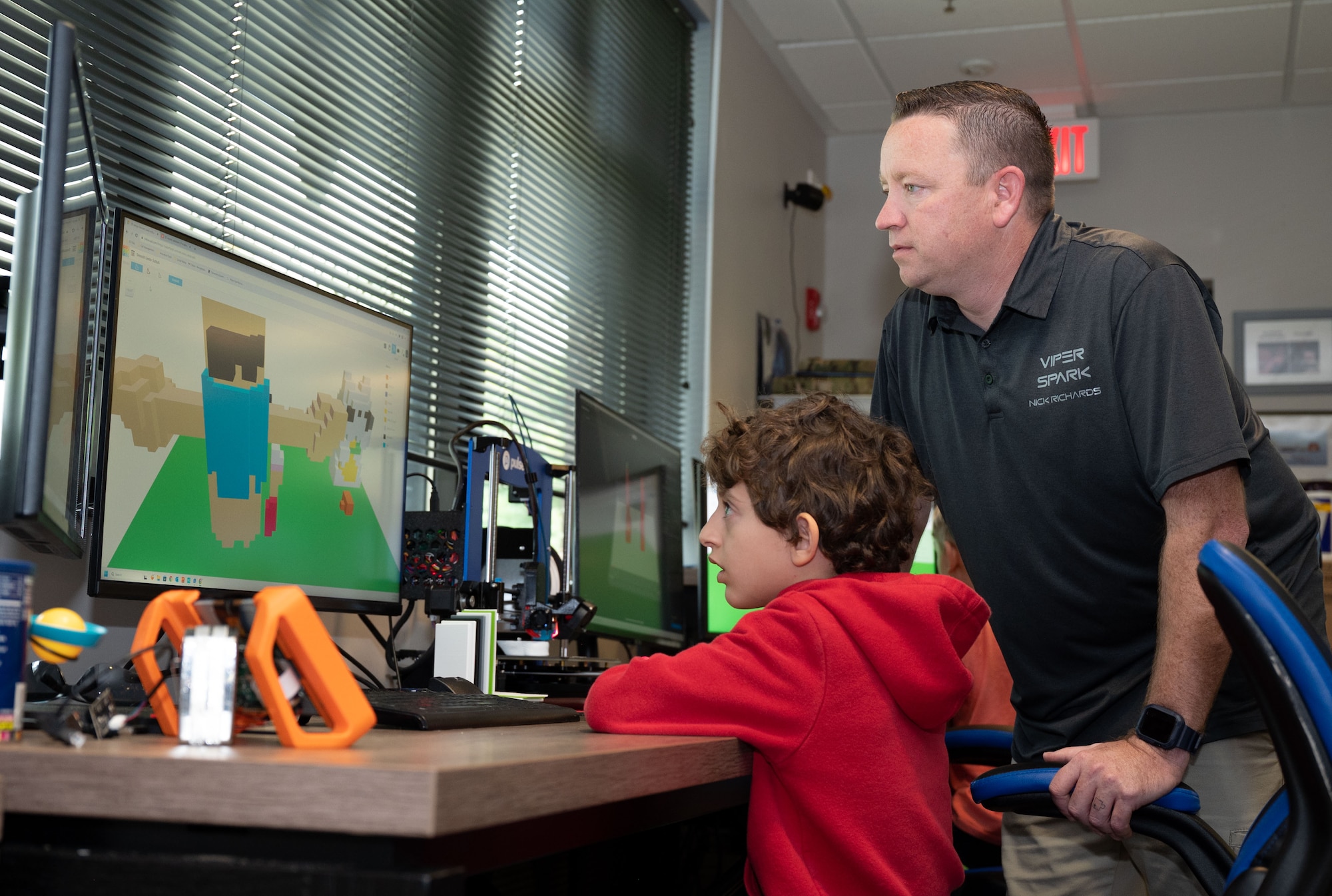 A man watches a computer screen standing behind a child sitting in front of the computer at a desk.