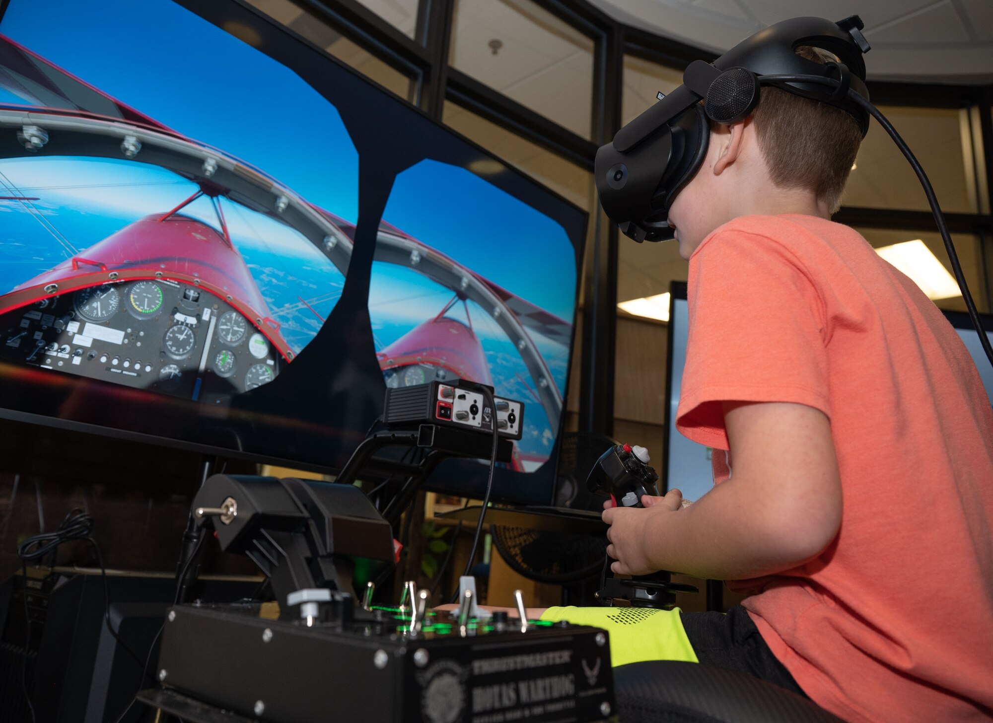 A child wearing virtual reality goggles holds a joystick on a flight simulator while watching two screens.