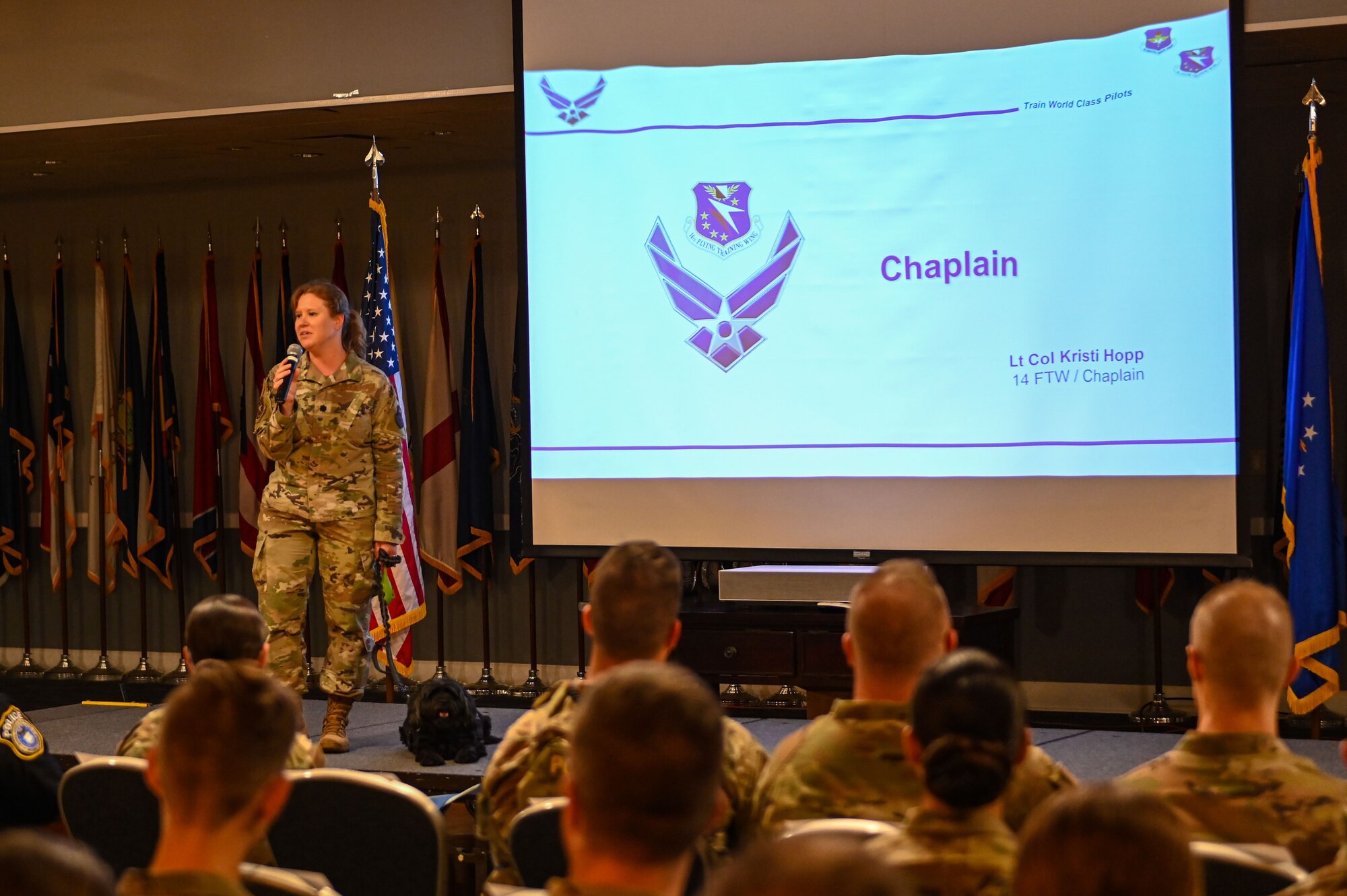 Lt Col. Kristi Hopp, 14th Flying Training Wing Head Chaplain grips Monk’s leash as she gives a briefing on becoming better every day at the Columbus Events Center July 21, 2023. Monk laid down on the stage next to Lt Col. Hopp as she finished her speech. (U.S. Air Force photo by Airman 1st Class Jessica Blocher)