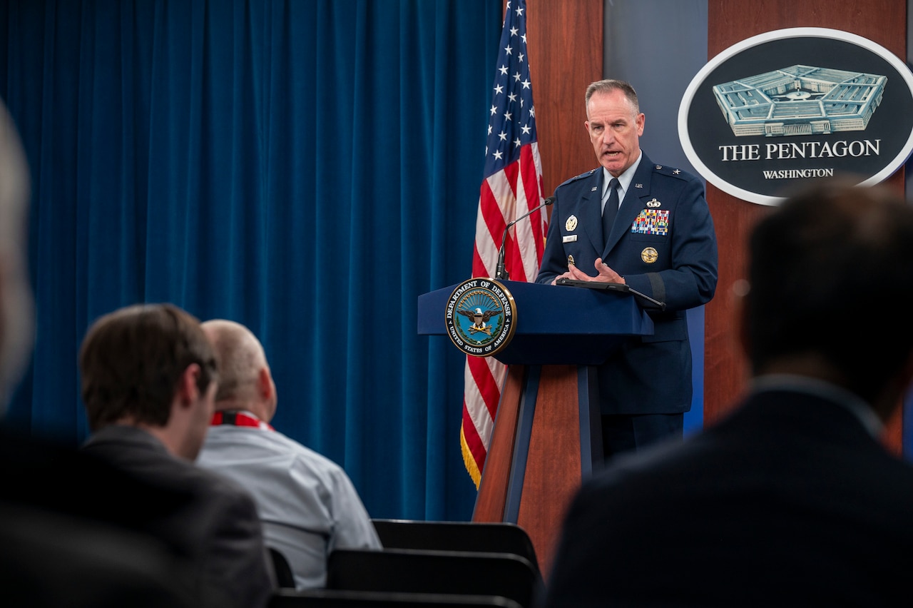 A person in uniform, standing at a lectern, speaks with members in an audience.