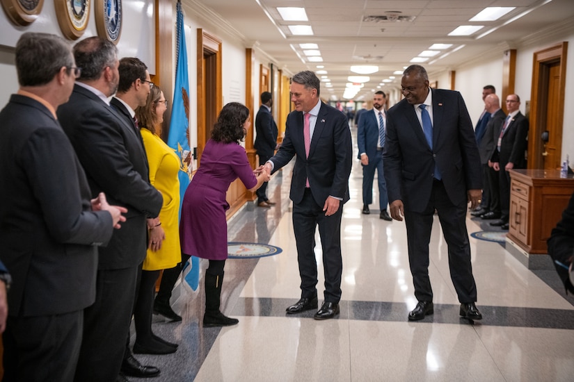 A man shakes hands with woman in a hallway as others stand around.