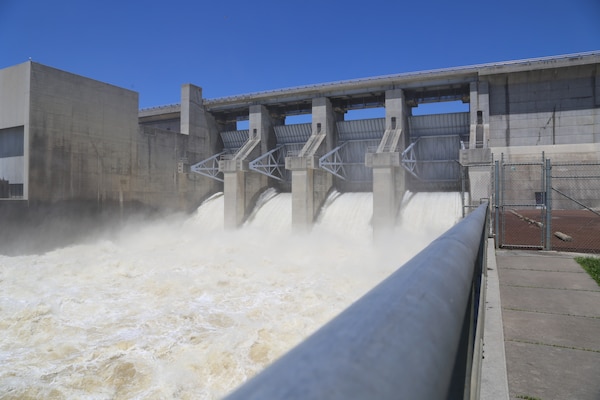 Water flows out of a dam with a concrete structure and blue sky in the background.