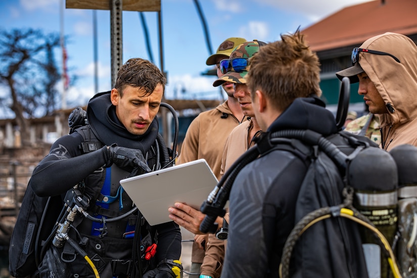 Service members wearing dive gear gather, as one points to a tablet.